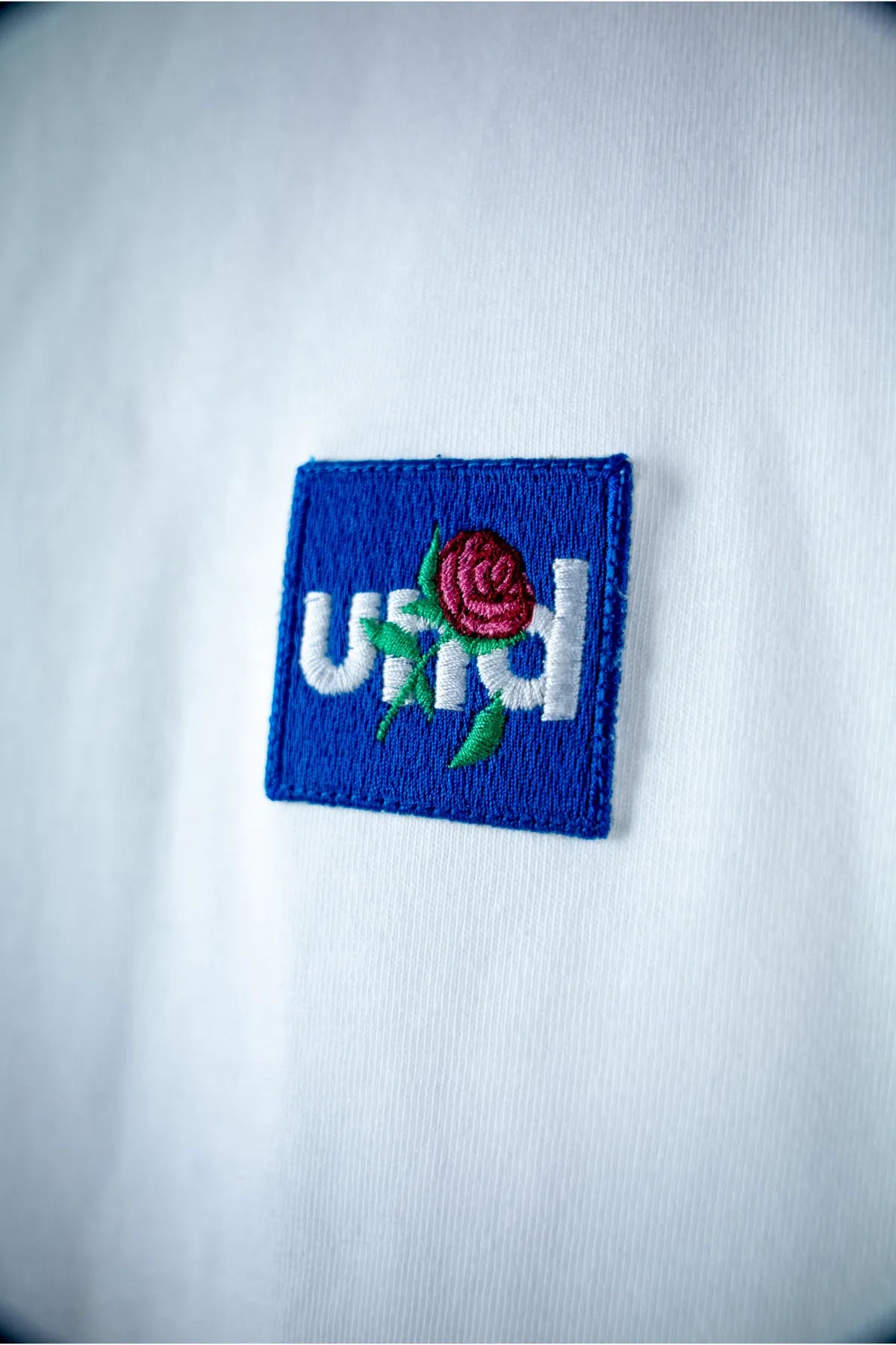 Und white T-shirt with "Und Rose Embroidery in Blue Badge"